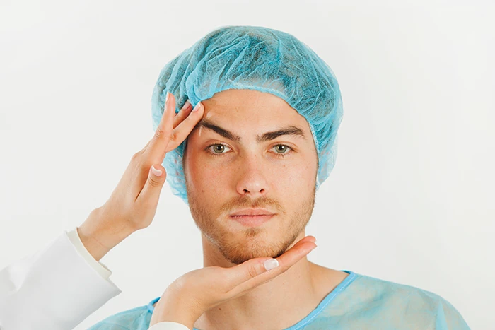 A cosmetic surgeon lifting a man’s eyebrow who is wearing a patient cap